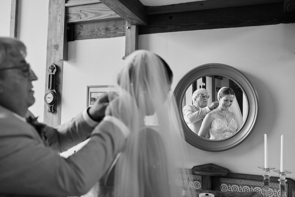 Black and white photo of the bride having her necklace fastened beside a round mirror