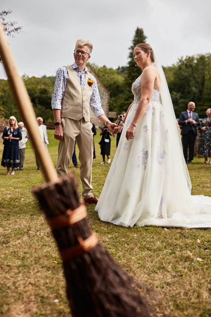 Bride and groom on the lawn with a broomstick in the foreground, ready to jump the broom