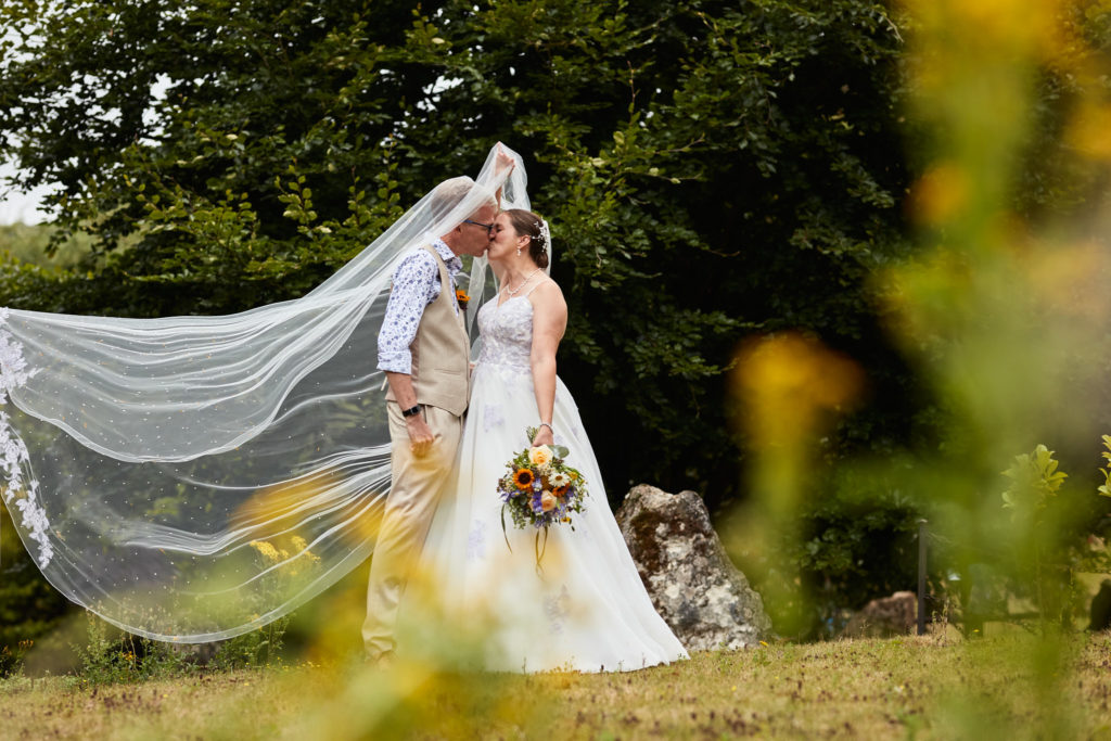 Bride and groom kissing as her veil blows in the wind, seen through flowers