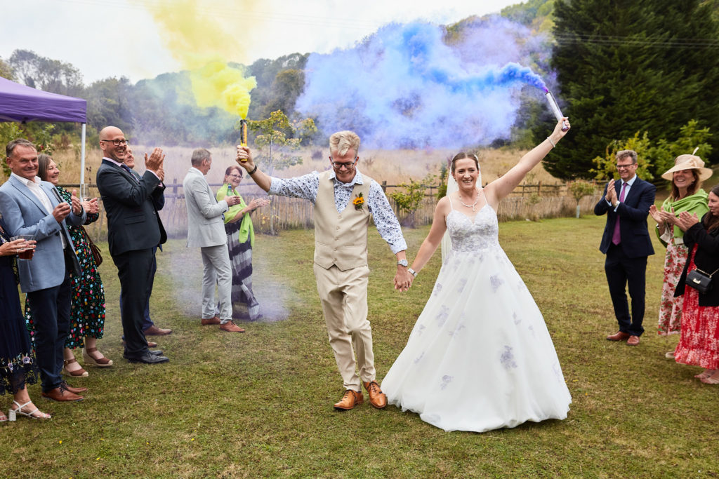 Bride and groom holding yellow and blue smoke flares as guests watch