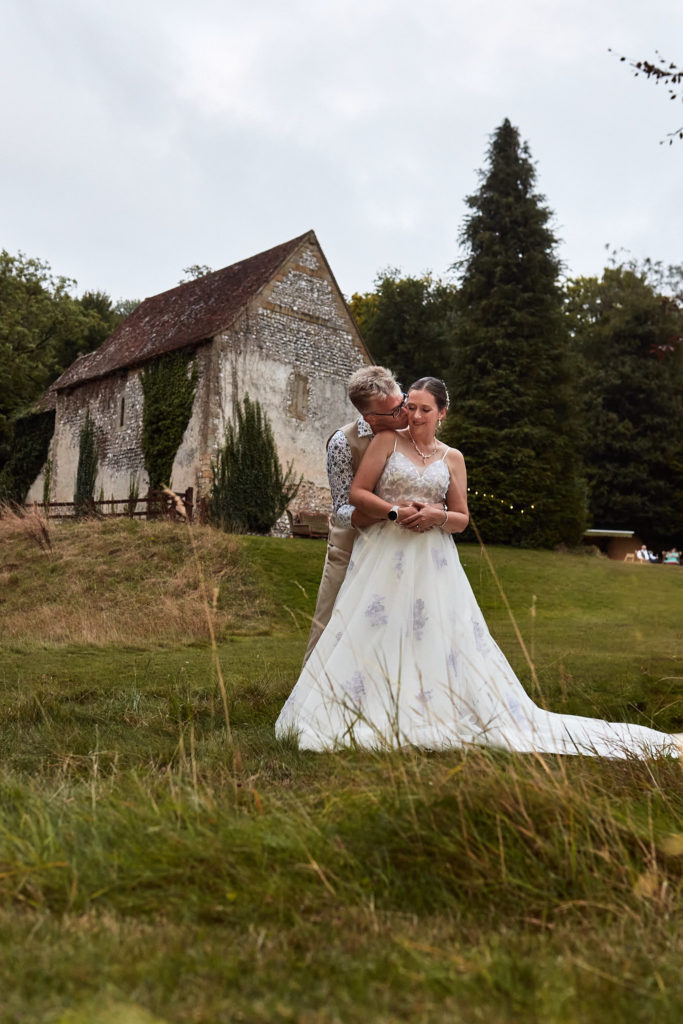 Groom embracing the bride from behind with the flint church at Dode behind them