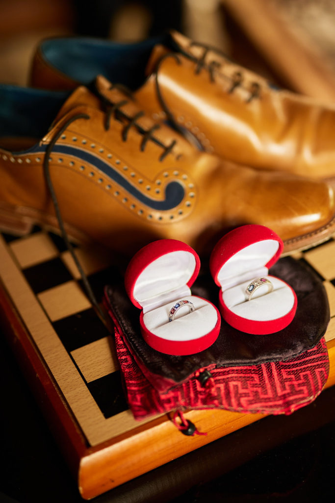 Wedding rings in red heart-shaped boxes beside tan brogues on a chessboard