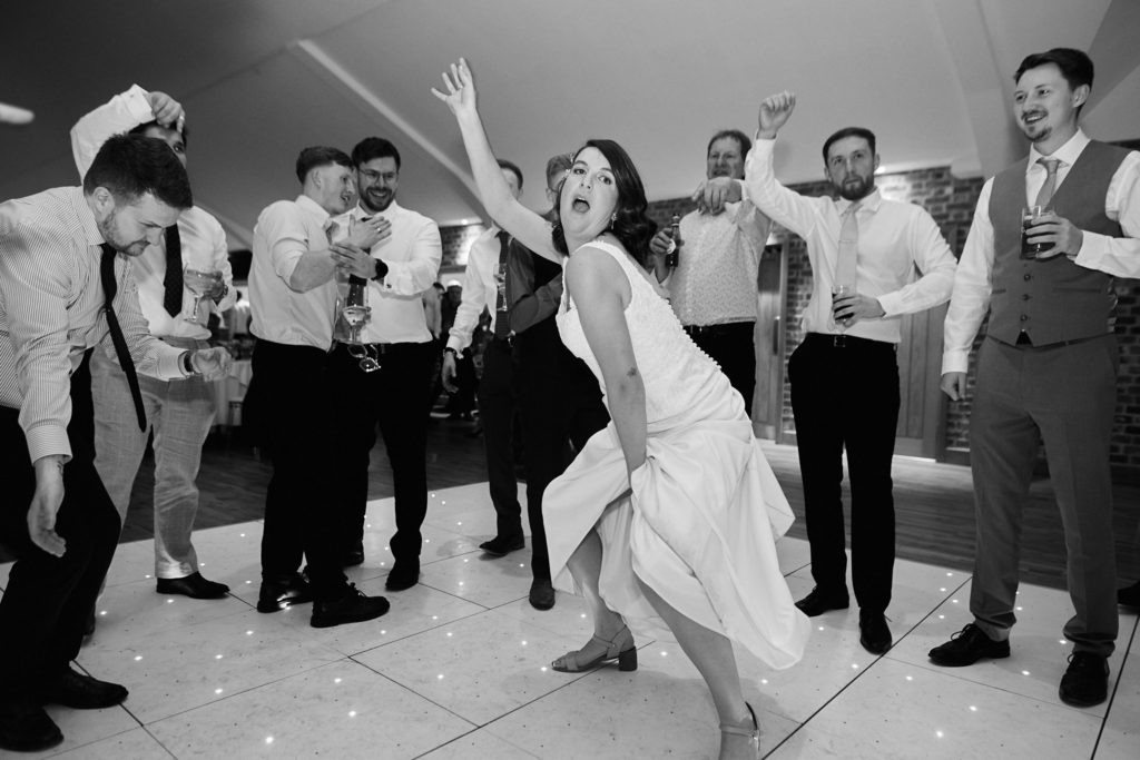 Black and white photo of the bride dancing with her arm in the air, surrounded by guests