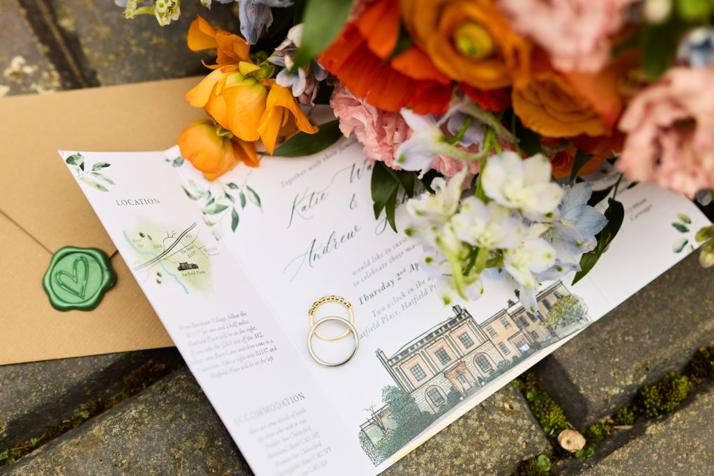 Wedding invitation with a green wax seal and wedding rings beside orange flowers