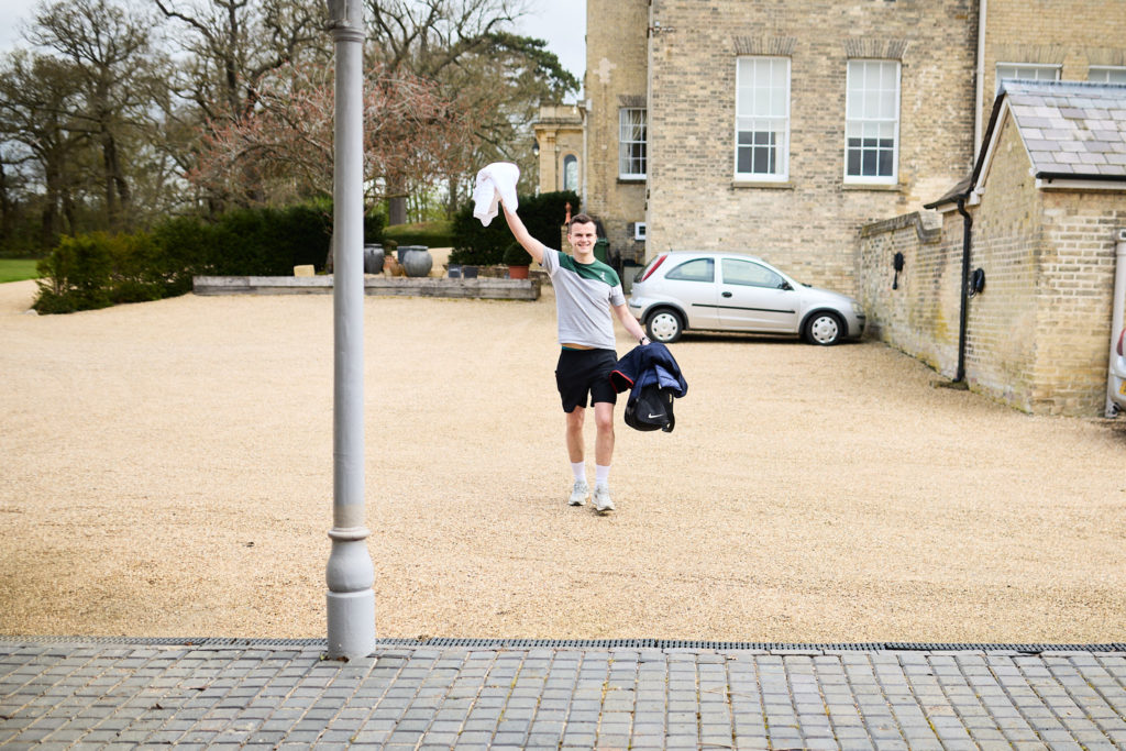 Man in a football shirt waving as he crosses the gravel courtyard carrying his suit