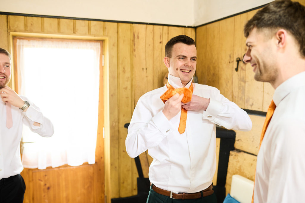 Groom laughing as he puts on his orange tie with a groomsman