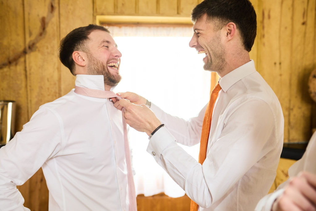 Groomsman adjusting the groom's collar as they both laugh