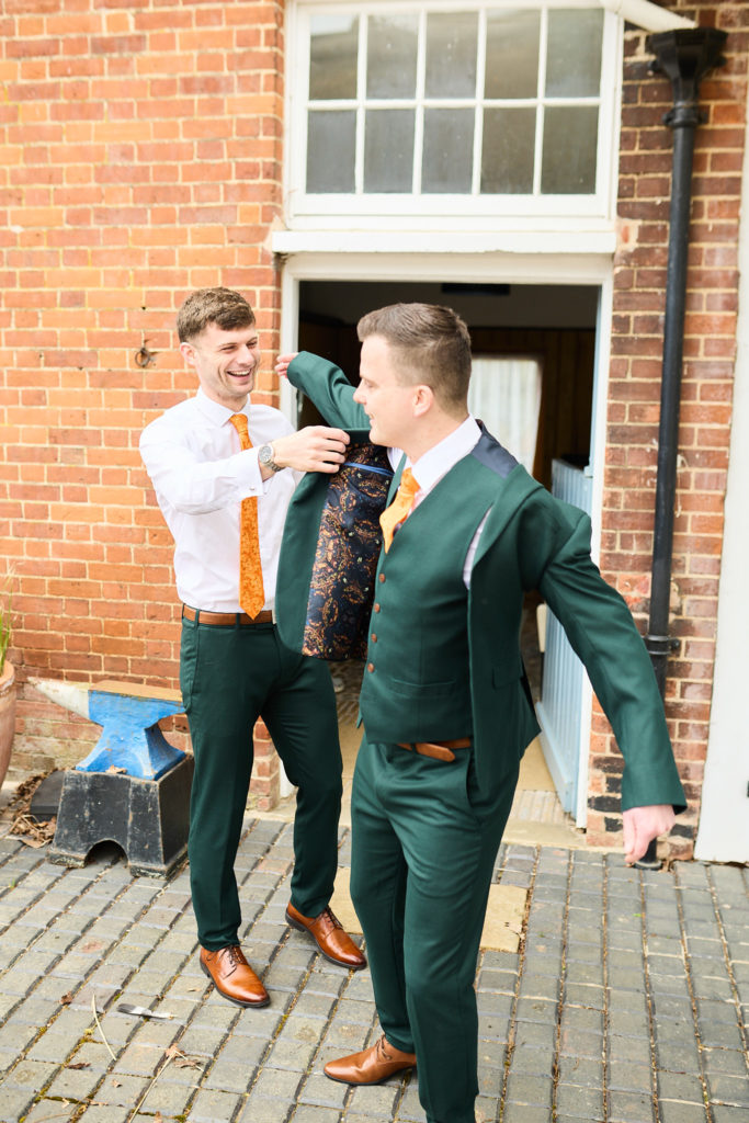 Groomsman helping the groom into his dark green suit jacket