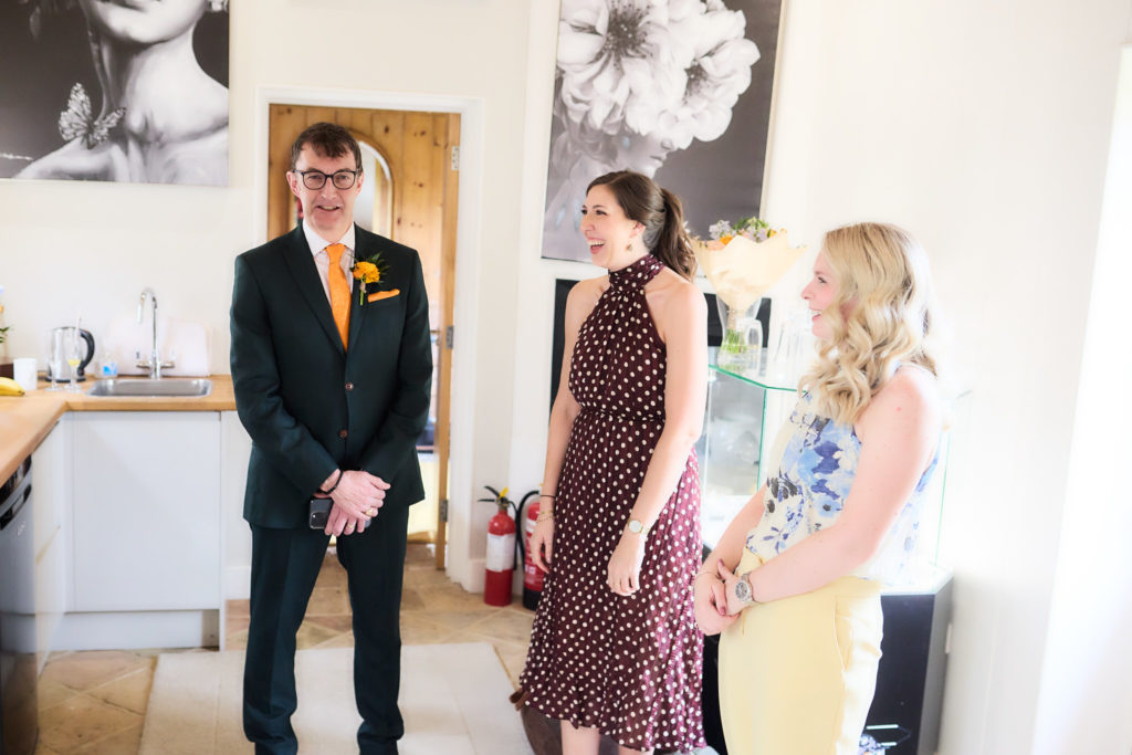 Groom in a dark green suit greeting two women guests indoors