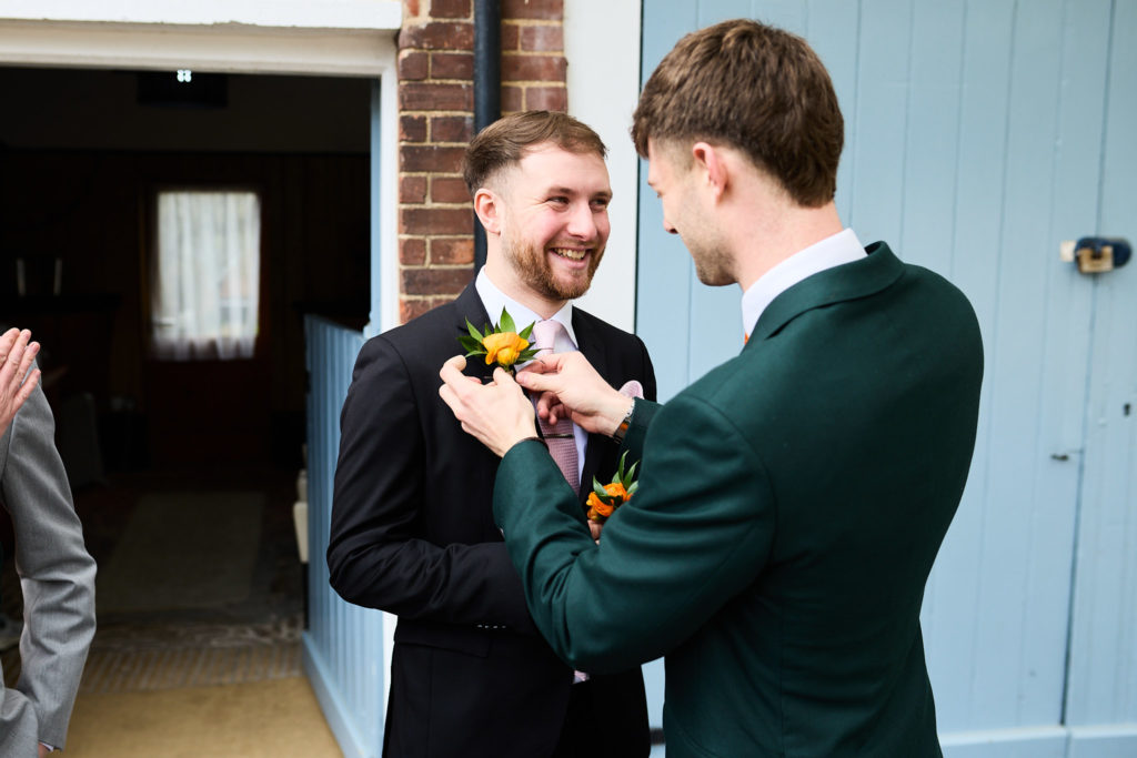 Groom pinning a buttonhole on a groomsman beside a pale blue door