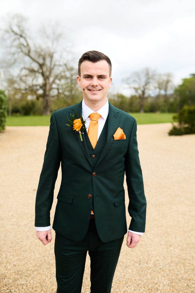 Groom smiling in a dark green suit with an orange tie and buttonhole