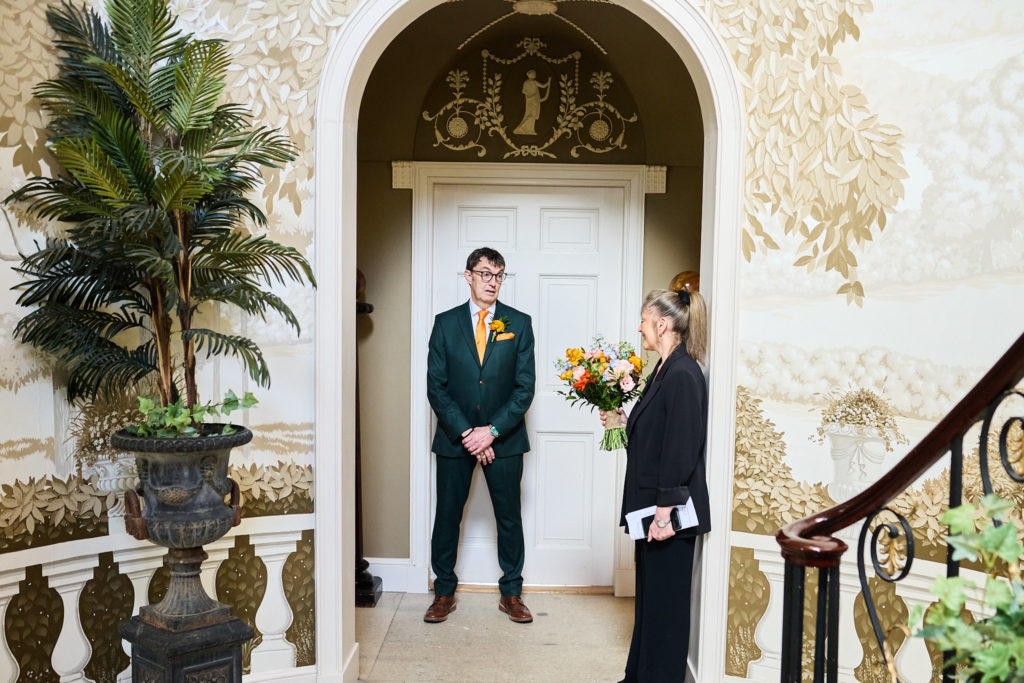 Groom in a green suit waiting in an arched doorway beside a woman holding the bouquet