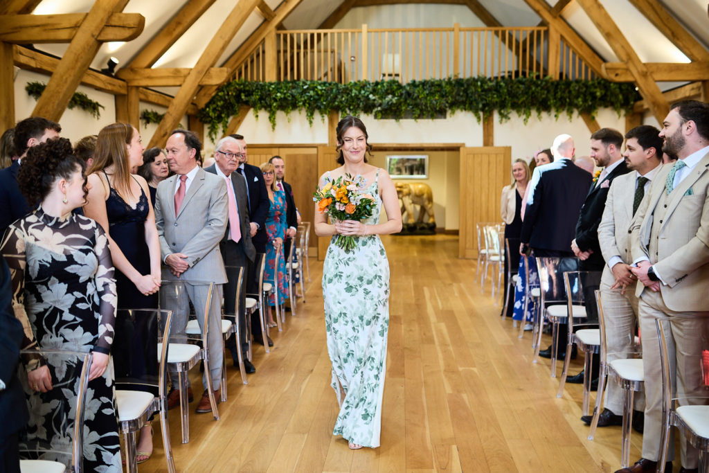 Bridesmaid in a blue floral dress walking down the aisle in the oak barn