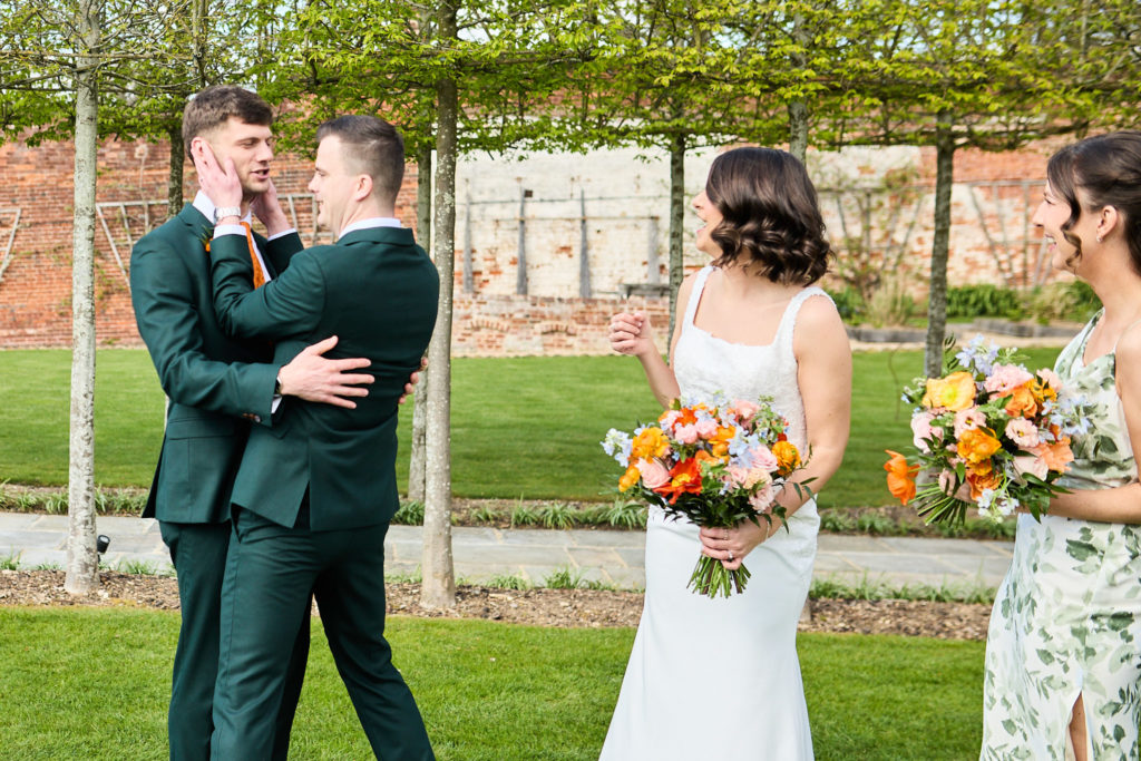 Groom hugging a groomsman as the bride and a bridesmaid look on with their bouquets