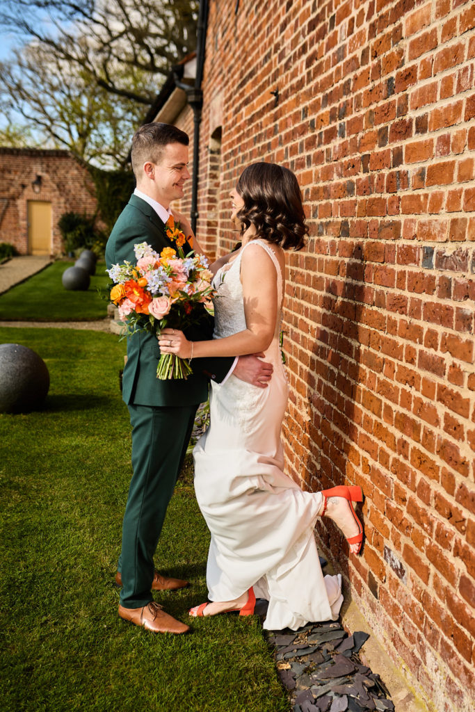 Bride kicking up her orange shoe as she leans into the groom against a red-brick wall at Hatfield Place, Essex