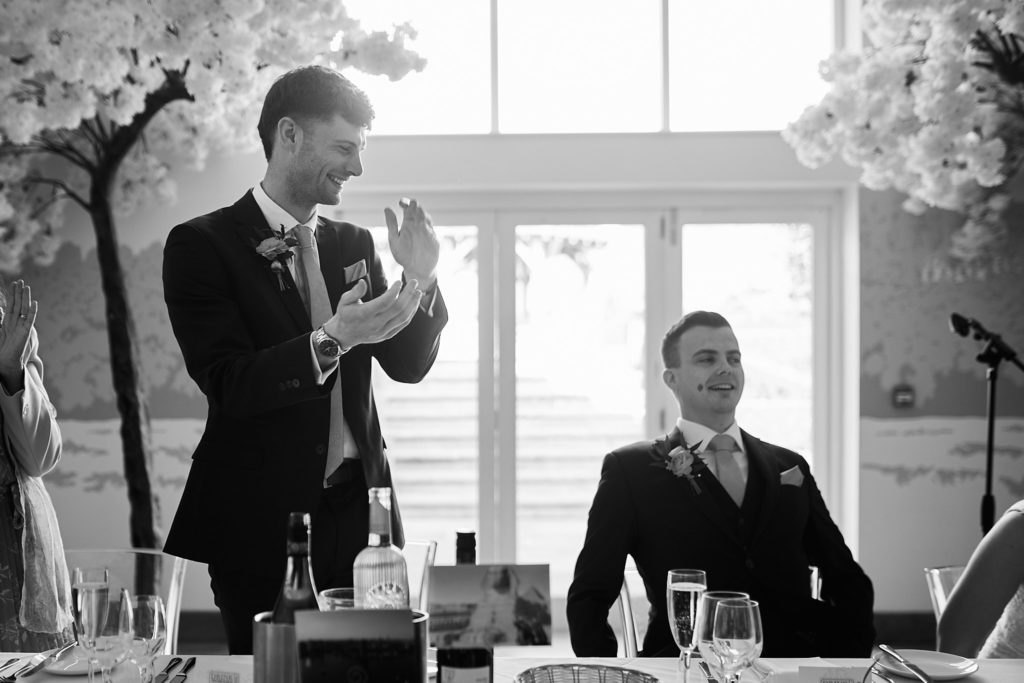 Black and white photo of a man applauding during the speeches as the groom looks on