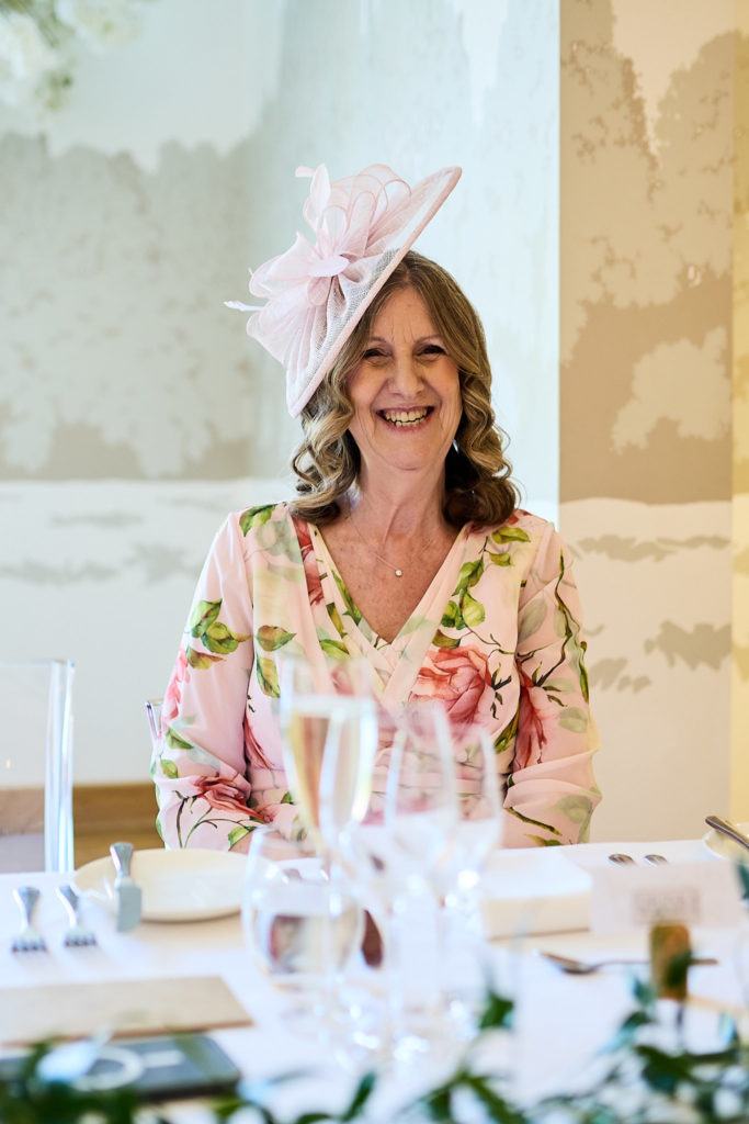 Woman in a pink fascinator and floral dress smiling at the table
