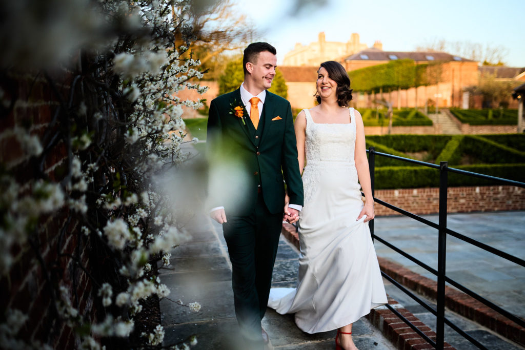 Bride and groom walking hand in hand past blossom in the gardens of Hatfield Place