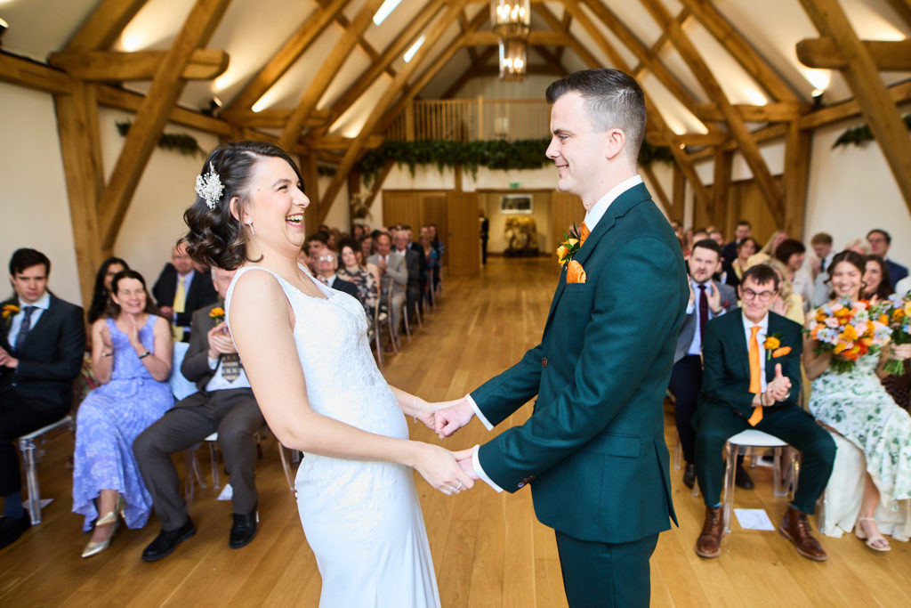 Bride and groom holding hands and laughing during their ceremony in the oak barn at Hatfield Place
