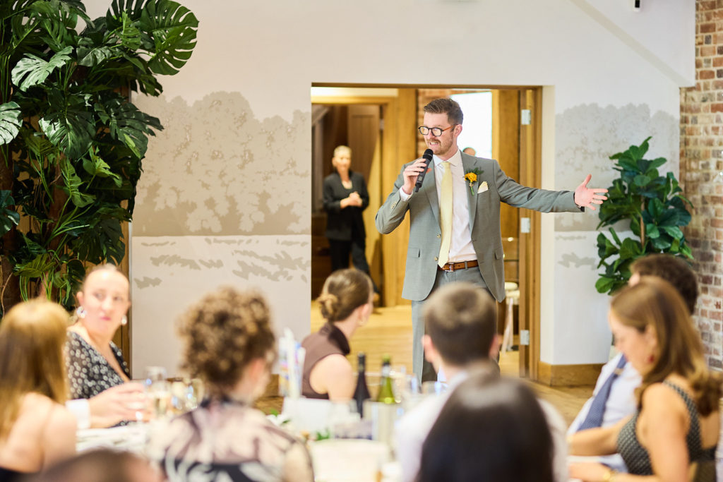 Man in a grey suit entering the room with his arms outstretched as guests look on