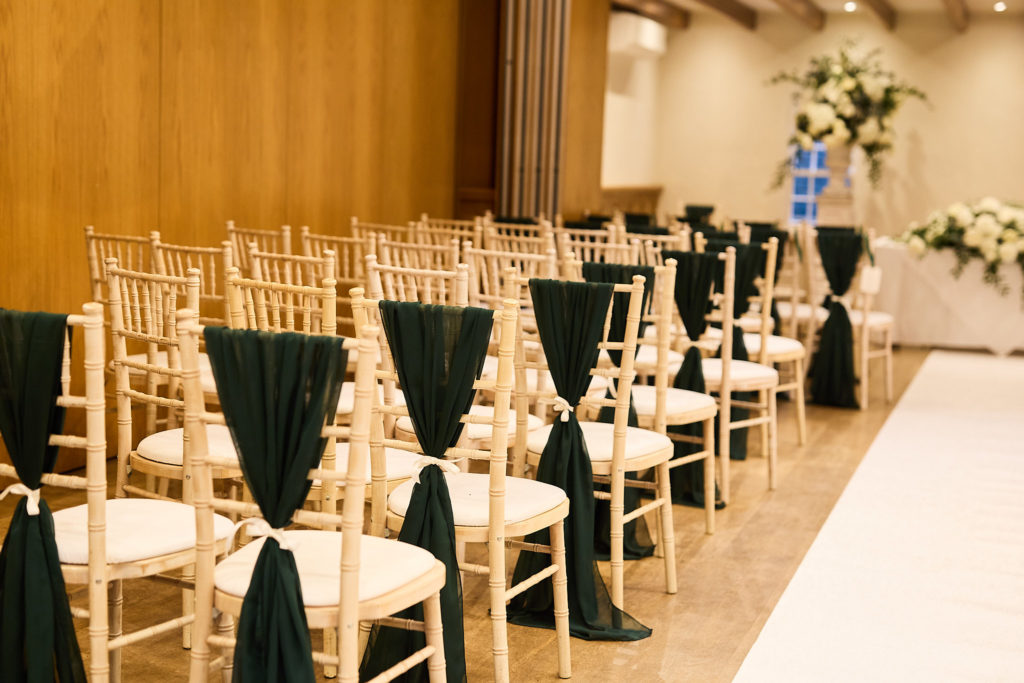 Rows of ceremony chairs with dark green sashes beside a white aisle