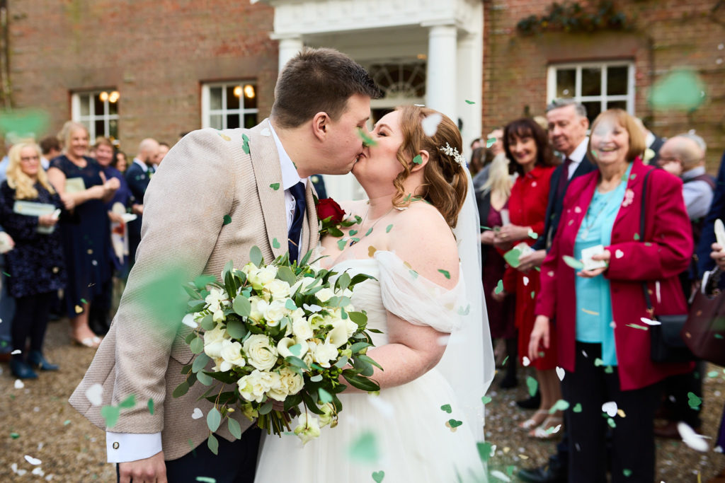 Bride and groom kissing as guests throw confetti at Mulberry House, Essex