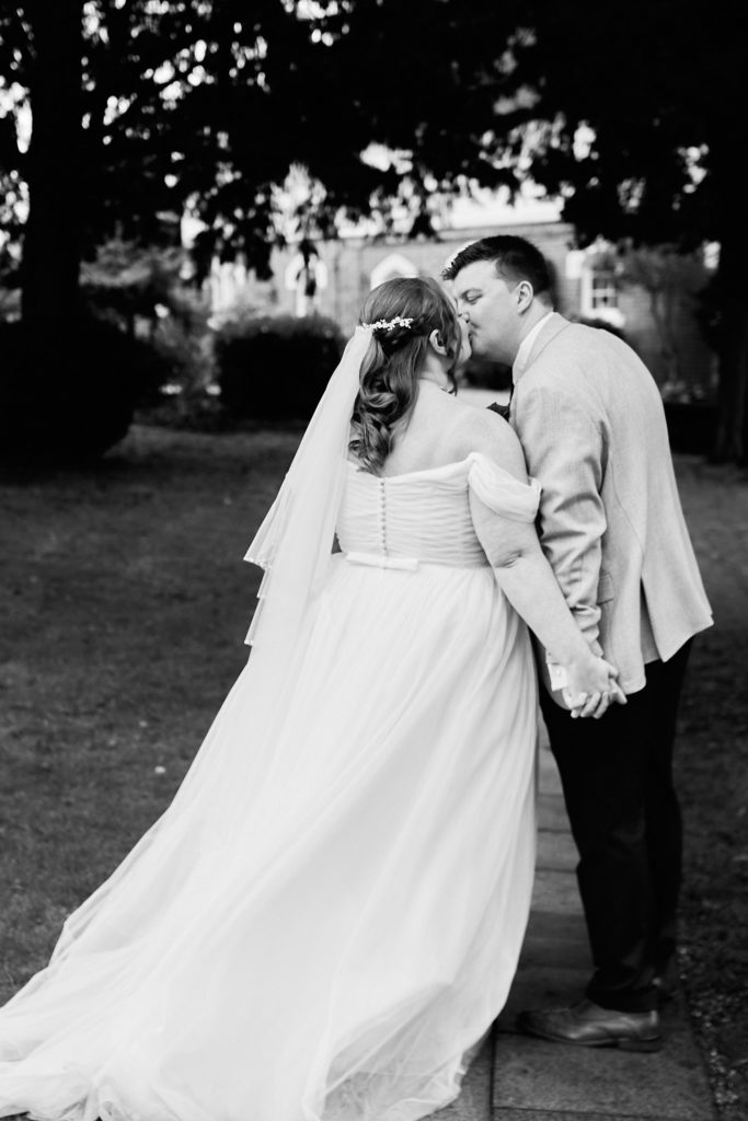 Black and white photo of the groom kissing the bride as they hold hands in the gardens of Mulberry House