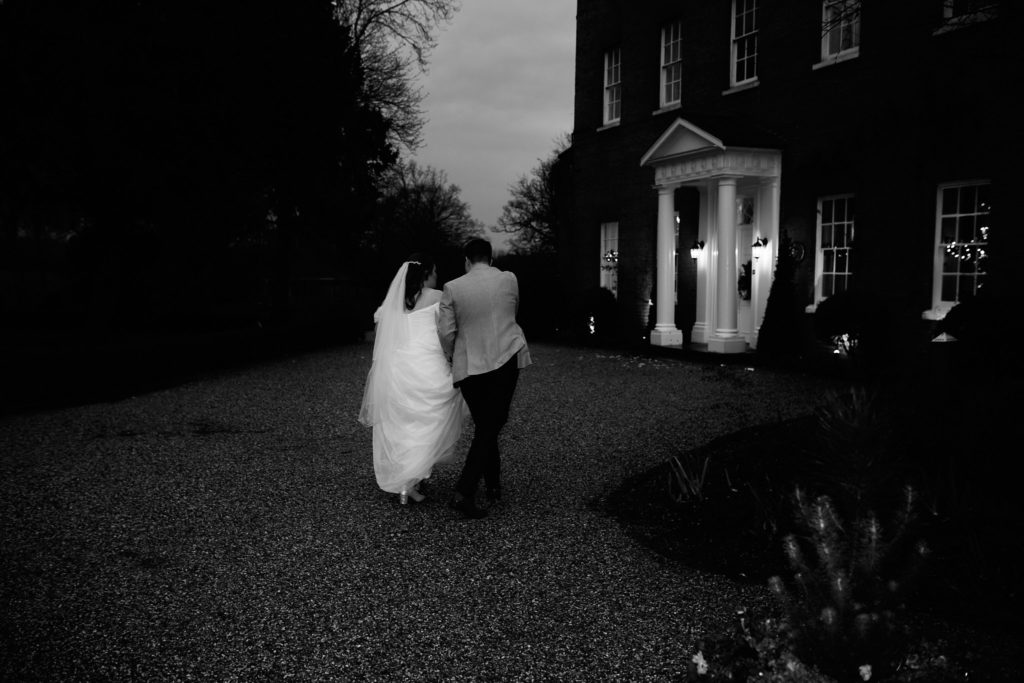 Black and white photo of the bride and groom walking towards Mulberry House at dusk