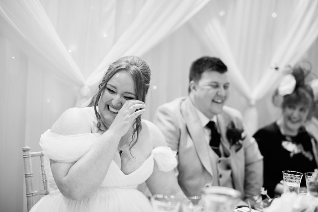 Black and white photo of the bride wiping away a tear beside the groom during the speeches