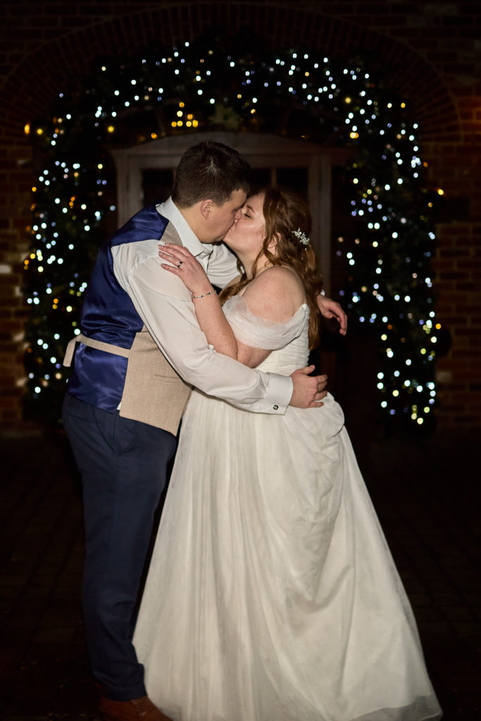 Bride and groom kissing at night in front of an arch of fairy lights at Mulberry House