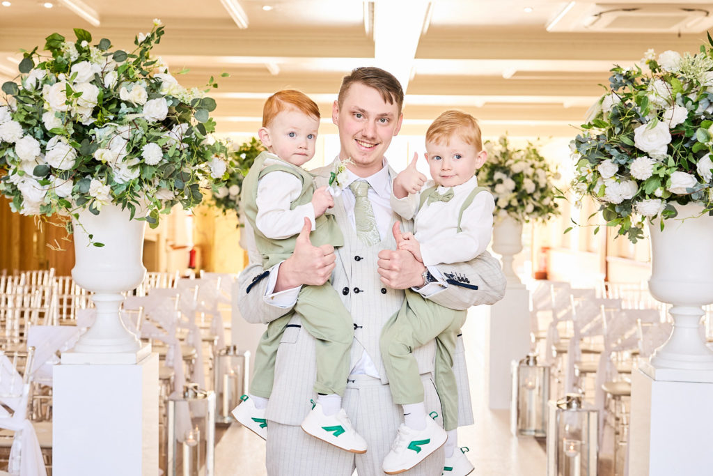 Groom holding two toddlers in matching cream outfits in the ceremony room
