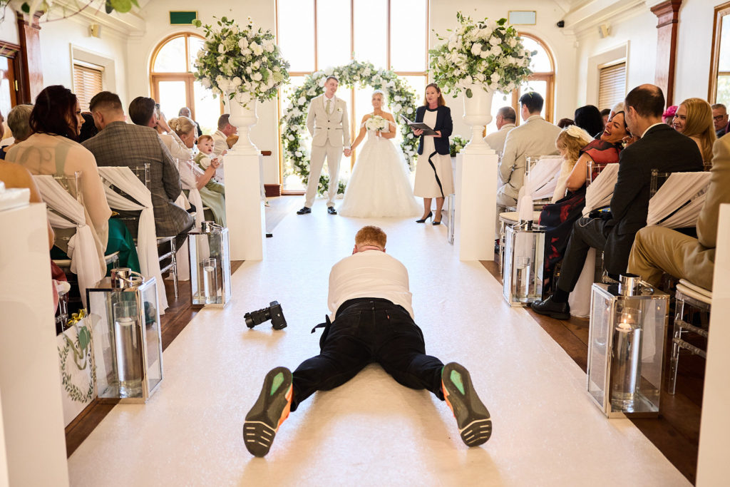 Groomsman lying face down in the aisle as guests look on before the ceremony