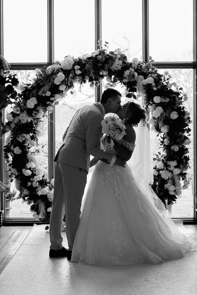 Black and white photo of the bride and groom kissing under a floral arch