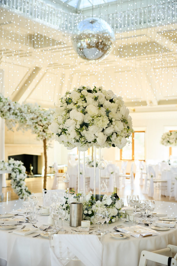 Round table set with a tall white floral centrepiece under fairy lights and a mirror ball