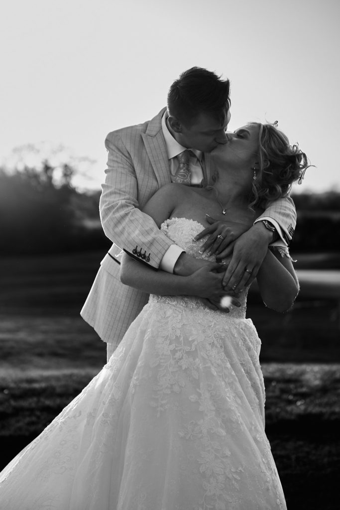 Black and white photo of the groom kissing the bride as she wraps her arms round his neck