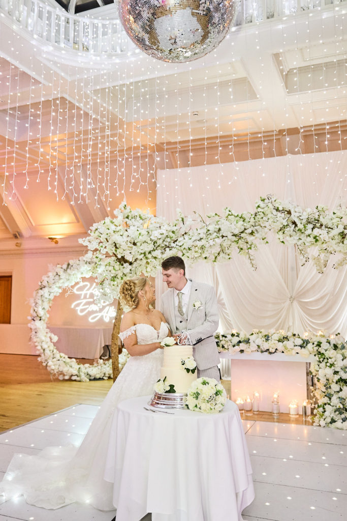 Bride and groom beside their wedding cake in front of a floral arch