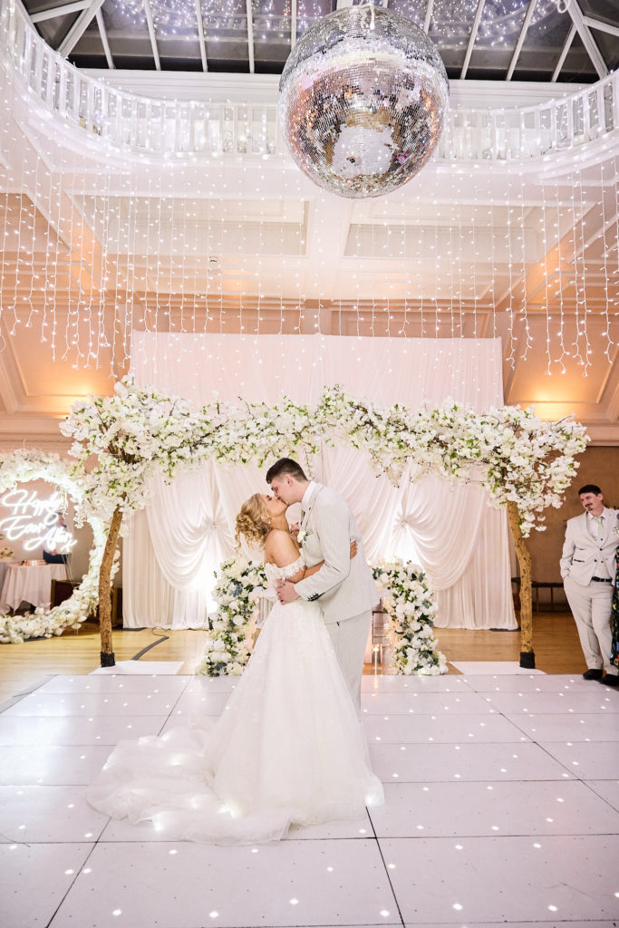Bride and groom kissing on a white dance floor under a mirror ball and floral arch
