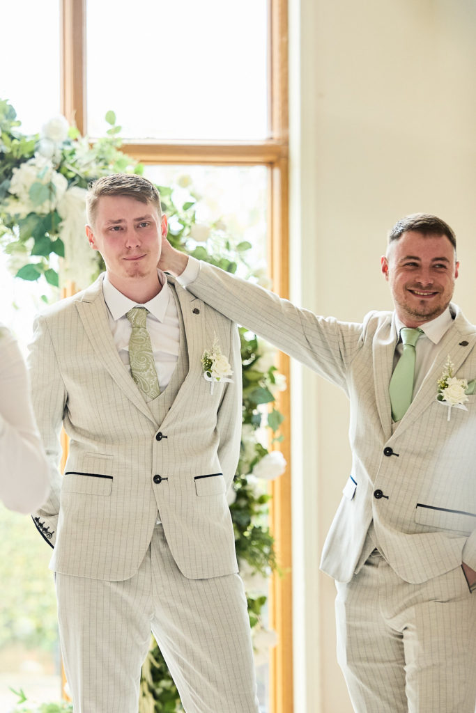 Groom and best man in cream suits joking beside a window