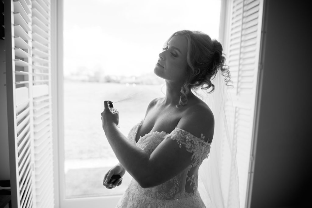 Black and white photo of the bride spraying perfume beside a window
