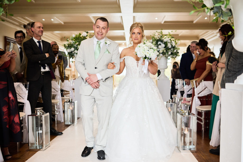 Bride walking down the aisle arm in arm with a man in a cream suit