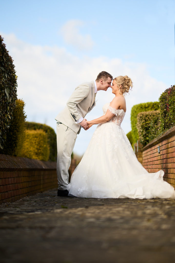 Bride and groom leaning in to kiss on a path at Stock Brook Manor under a blue sky