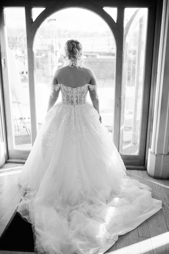 Black and white photo of the bride from behind at a window, showing the full skirt of her dress
