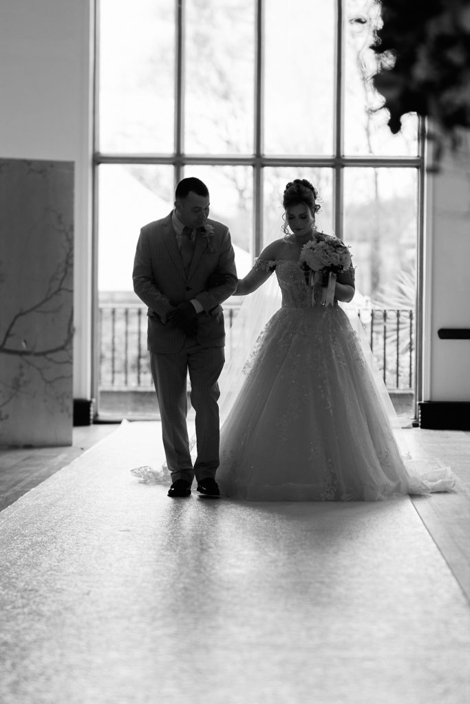 Black and white photo of the bride walking arm in arm with a man towards the ceremony