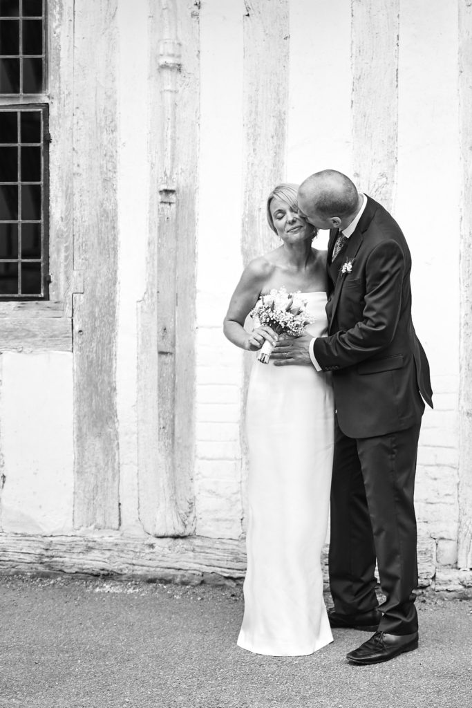 Black and white photo of the groom kissing the bride's cheek in front of a timber-framed house in Lavenham