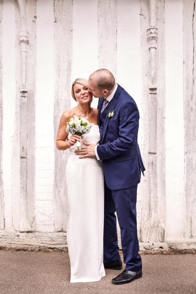 Bride laughing as the groom kisses her cheek beside a timbered wall in Lavenham