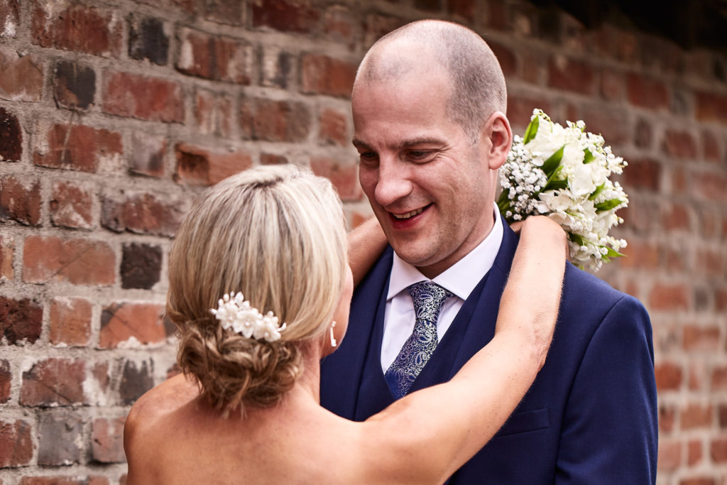 Groom smiling as the bride wraps her arms round his neck in front of a brick wall