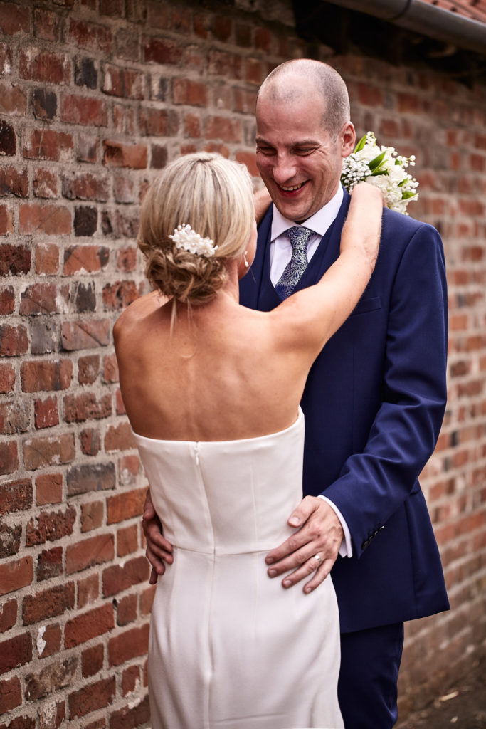Groom laughing as the bride embraces him against a red-brick wall in Lavenham