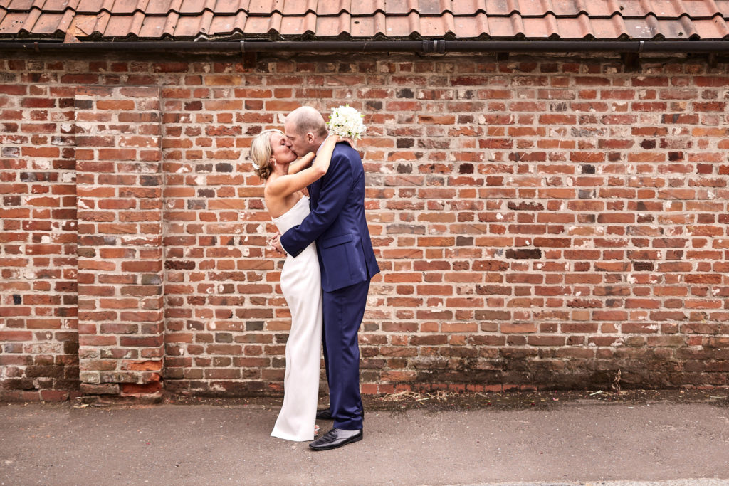 Bride and groom kissing in front of a long red-brick wall in Lavenham