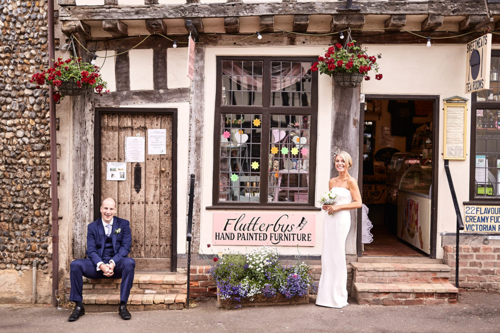 Groom sitting on the step of a timber-framed shop in Lavenham with the bride standing in the doorway