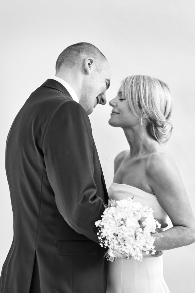 Black and white photo of the bride and groom nose to nose with her bouquet between them
